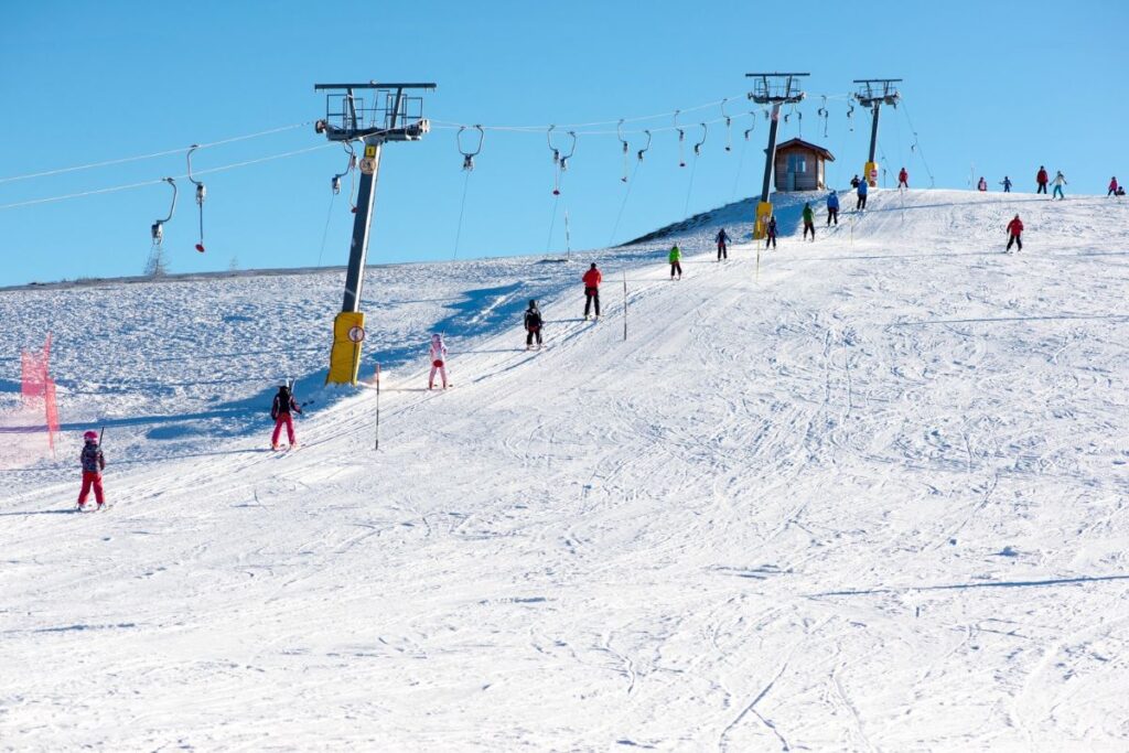 Beginner skiers on a wide gentle slope beside a surface lift with red-and-yellow safety padding