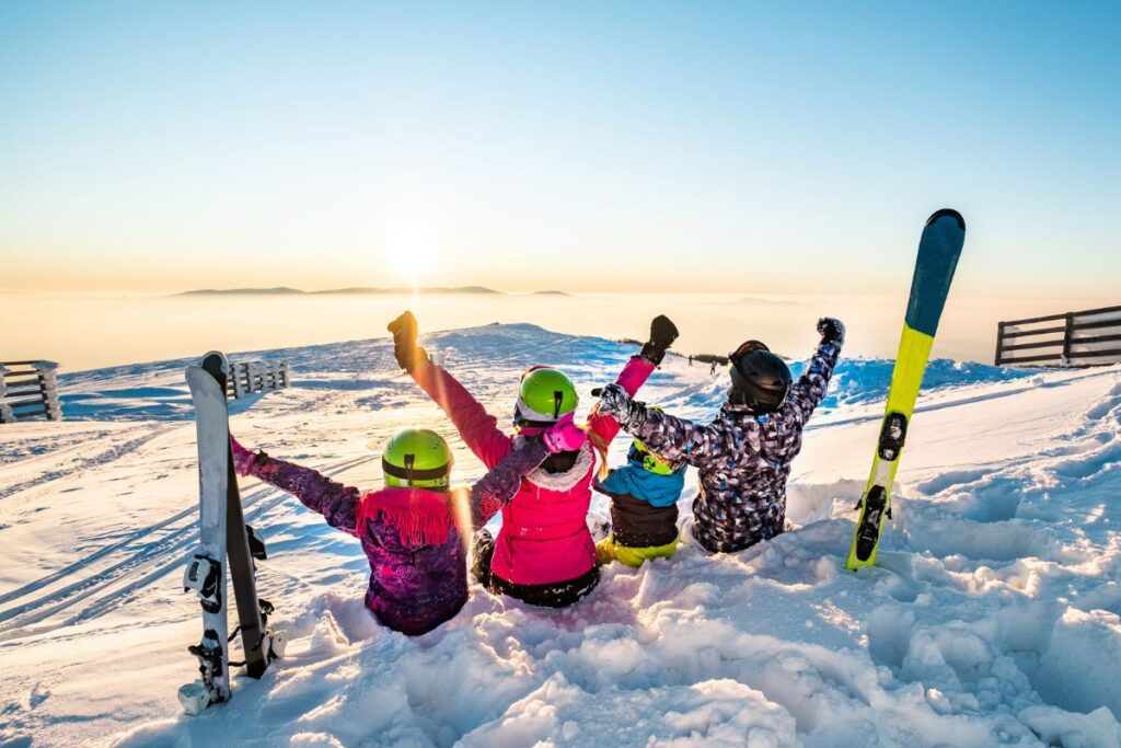 Two adults and two children in ski helmets sitting in the snow with arms raised, skis planted beside them